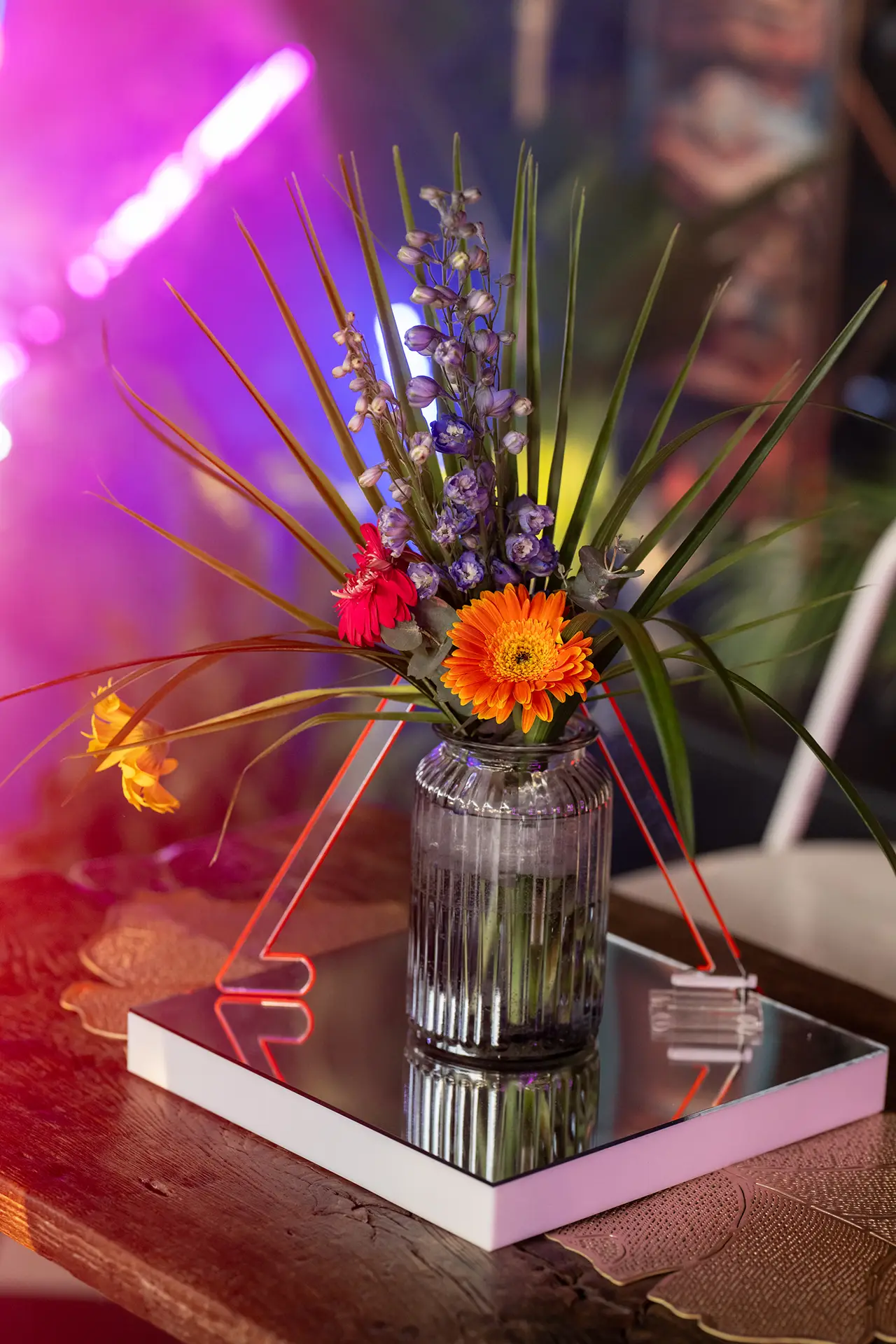 Glass jar vase with orange and red flowers, purple buds, and green leaves on a mirrored tray.